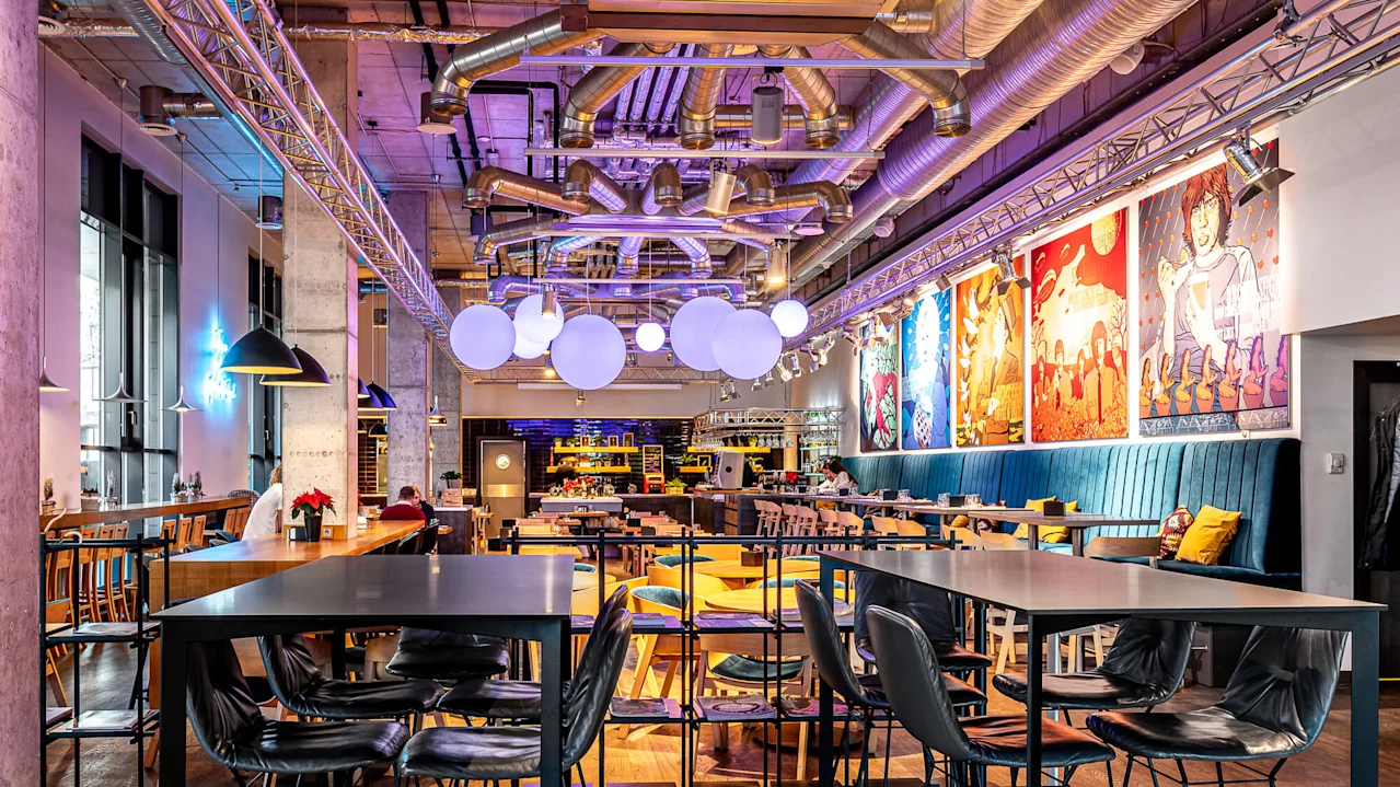 Modern dining space with chairs and tables, vibrant artwork on the walls, and large, spherical lights overhead. Industrial decor with exposed ducts and colorful ambiance.