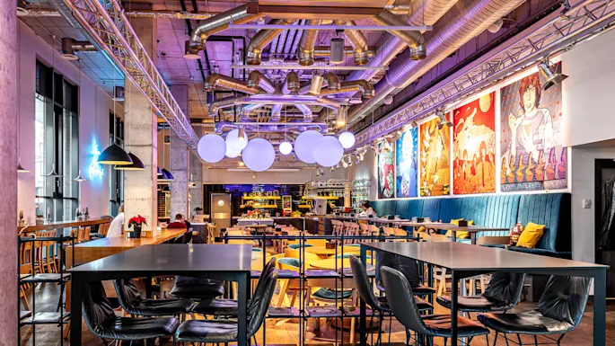 Moderne spisested med stoler og bord, fargerike kunstverk på veggene og store, sfæriske lamper over hodet. Industrielt interiør med eksponerte kanaler og fargerik atmosfære.