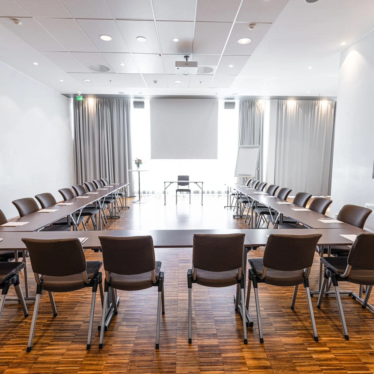 A U-shaped conference table with black chairs is set up in a bright room with wooden floors and framed artwork on the walls.