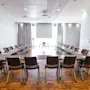 U-muotoinen konferenssipöytä, jossa on mustia tuoleja, on asetettu valoisaan huoneeseen, jossa on puulattia ja kehystettyjä taideteoksia seinillä.