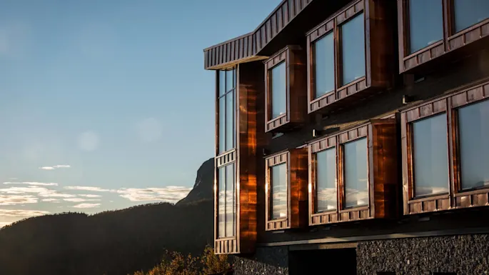 A modern building with large reflective windows, partially bathed in sunlight, stands against a backdrop of distant hills and a clear blue sky.
