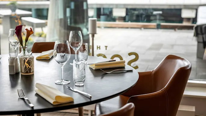 Round table set with neatly arranged glassware, utensils, and napkins; surrounded by brown chairs in a modern restaurant environment with large windows. Text on glass: "THE SSOS" (partially visible).