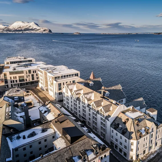 Luftfoto av snødekte bygninger ved vannkanten i Ålesund ved fjorden med fjell i bakgrunnen.