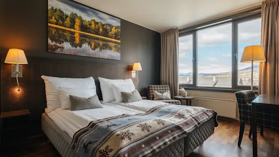 Ett mysigt hotellrum med en dubbelsäng, mönstrat överkast, en tavla på väggen och ett stort fönster med utsikt.