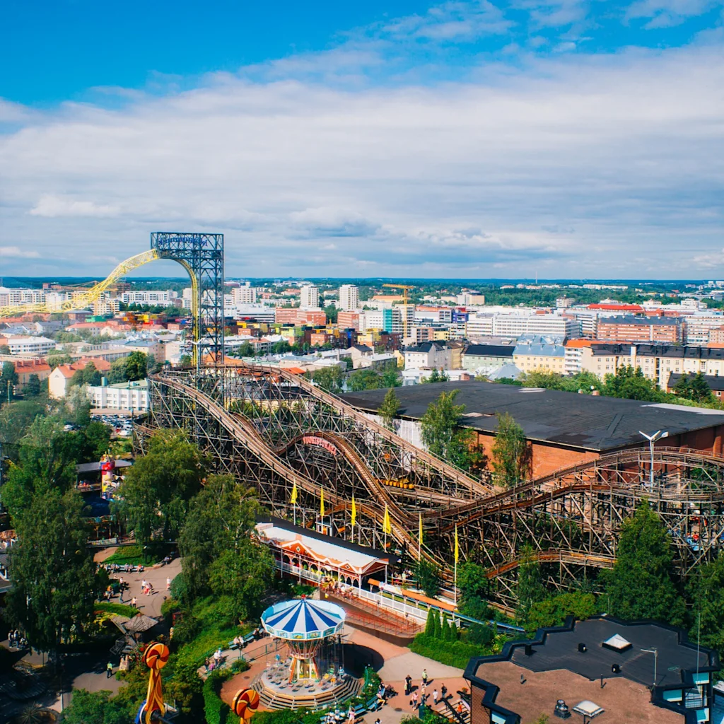 En luftfoto av Linnanmäki fornøyelsespark med berg-og-dal-baner, et høyt fritt fall-tårn og andre attraksjoner under en blå himmel.