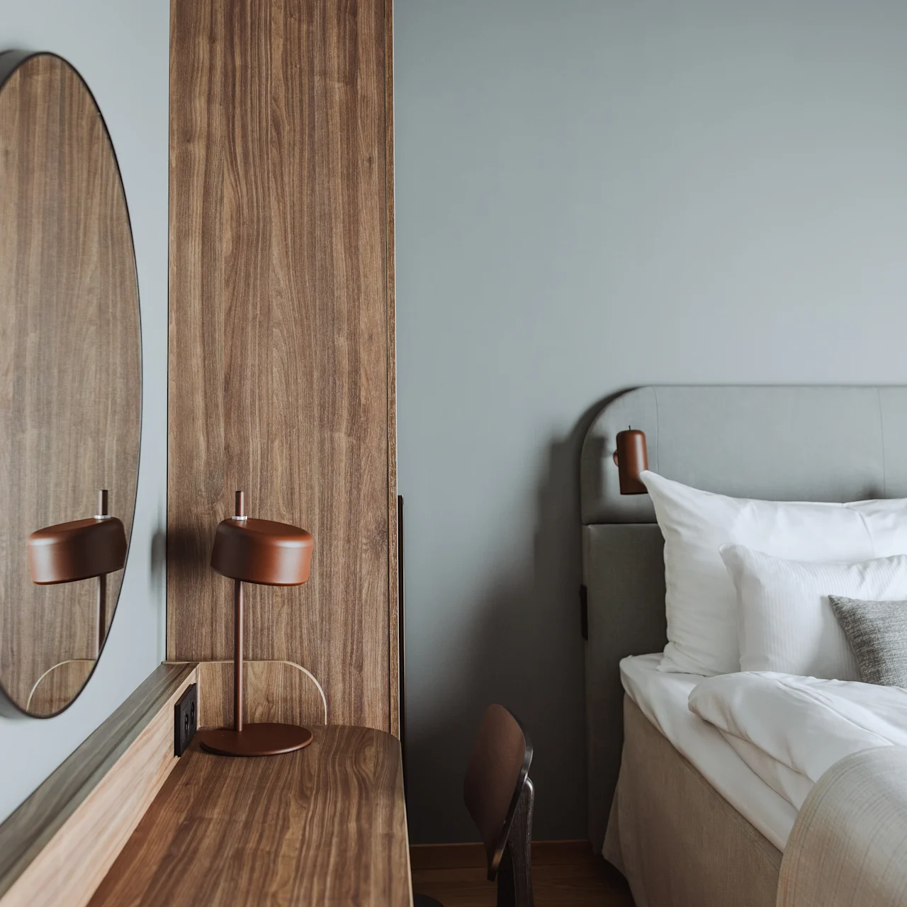 Rund spegel som reflekterar en minimalistisk lampa på ett träskrivbord. Modernt sovrum med en mjuk grå säng och vita sängkläder i närheten.