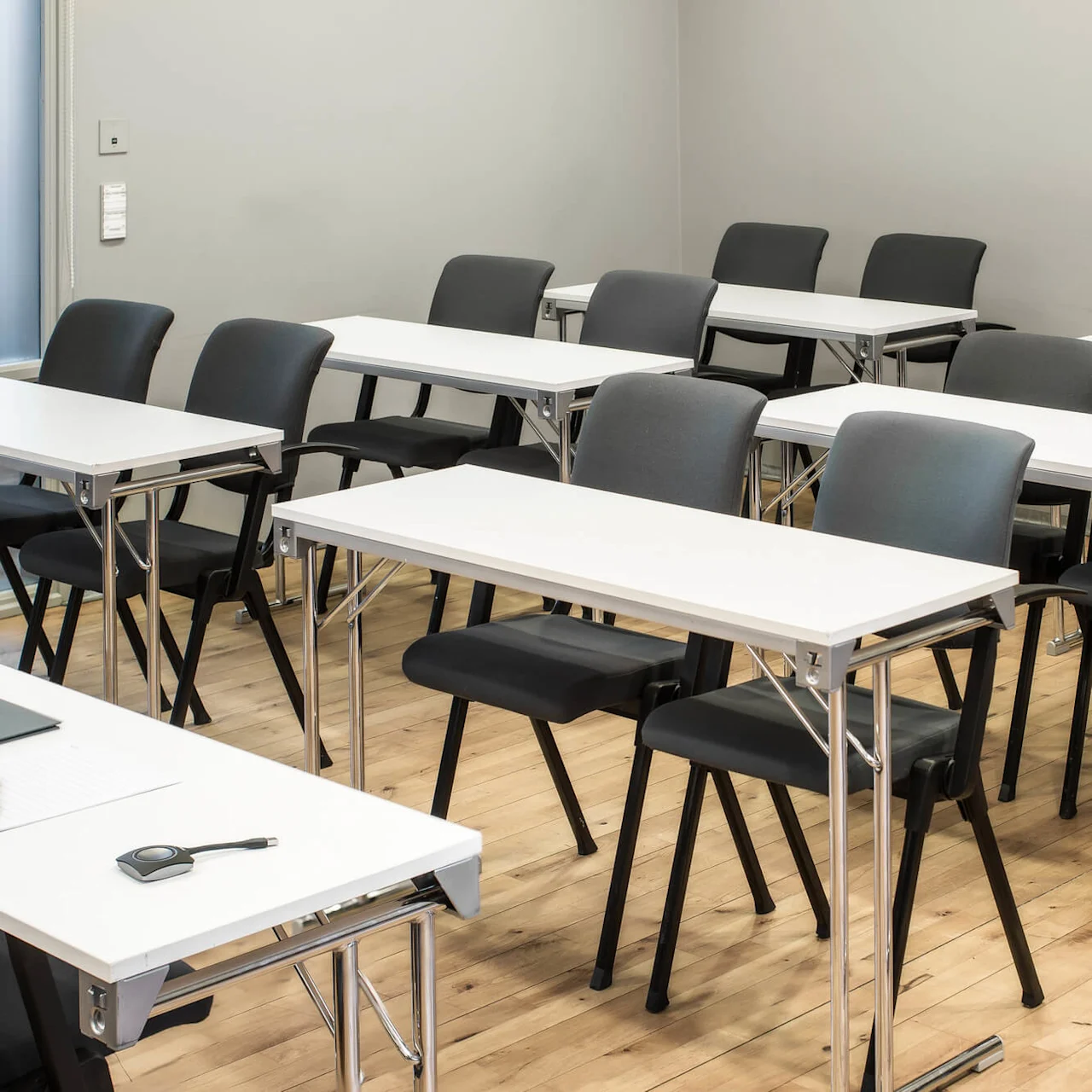 Desks and chairs are arranged in rows in a classroom setting with wooden flooring. A notebook, pen, and remote are on one desk.
