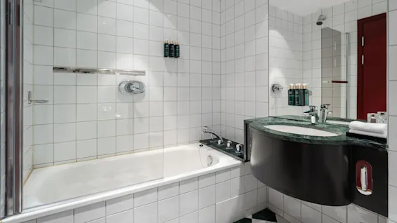 Badekar med krombeslag sidder ved siden af en flisevæg under skarpt lys, ved siden af en vask. Flasker er pænt arrangeret på hylden.