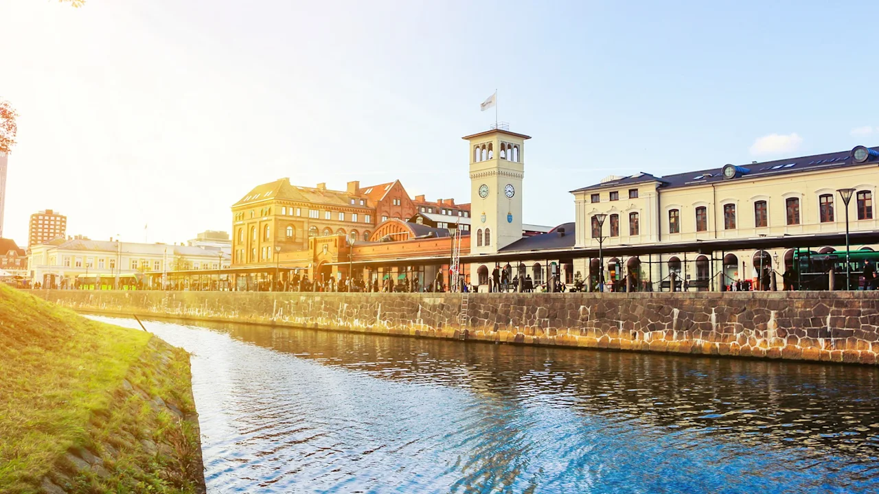 A canal flows alongside historic buildings with a clock tower, bathed in warm sunlight, reflecting a serene urban setting.