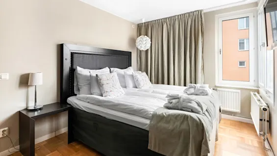 A neatly made bed with pillows, in a room with a window, curtains, a lamp, and a piece of wall art.