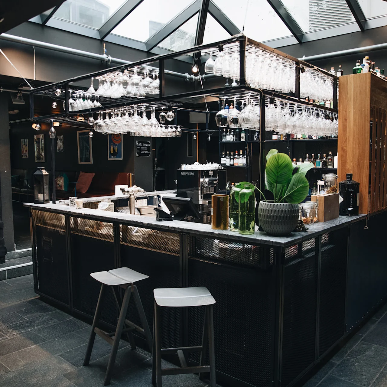 En moderne bar med hengende glass og to krakker, omgitt av svakt opplyste lounge-seter. Taket har takvinduer som forsterker den moderne atmosfæren.