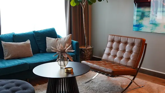 A blue sofa holds pillows beside a black round table with a vase, situated near a brown chair and a tall plant, under soft indoor lighting.
