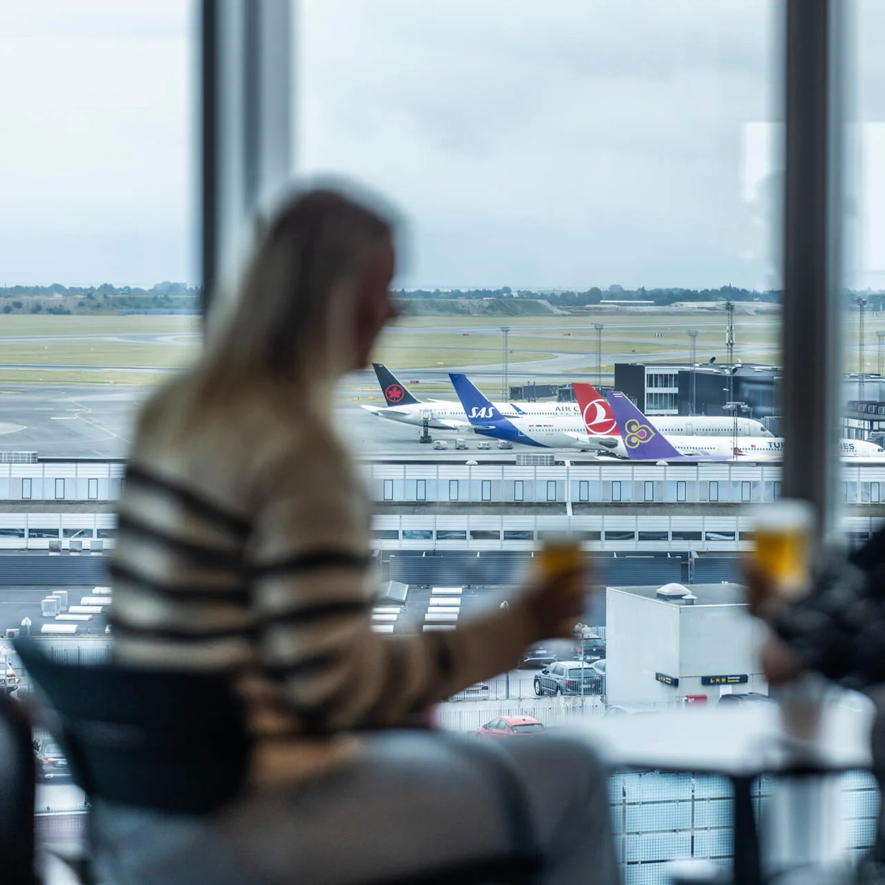 Utsikt over flyplassen med flere fly parkert ved terminalen, sett gjennom et stort vindu. To personer sitter ved et bord og nyter drikke mens de ser ut over rullebanen.