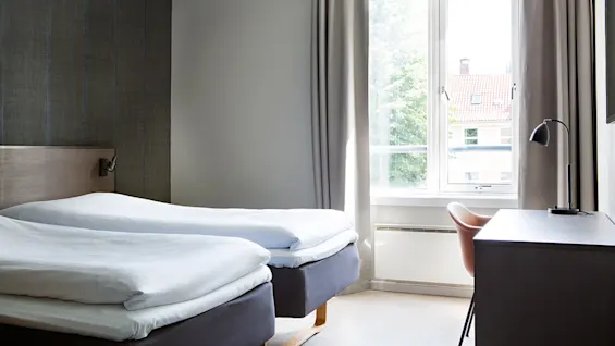 Senge er pænt redt med hvidt sengetøj i et hotelværelse. Et vindue slipper naturligt lys ind, hvilket oplyser et skrivebord med lampe og stol. Neutrale toner dominerer.