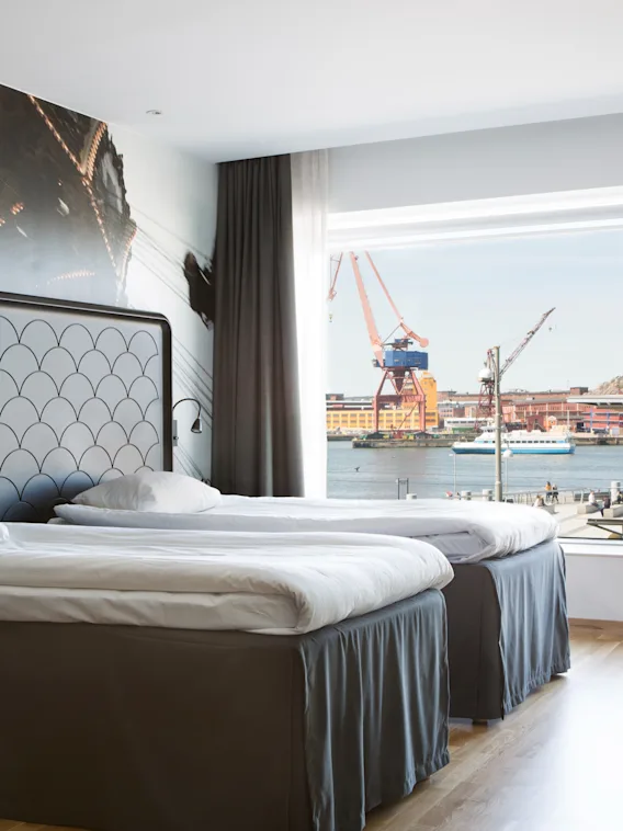 Et hotellrom med to senger og et stort vindu som gir utsikt over en travel havn med kraner og vann.