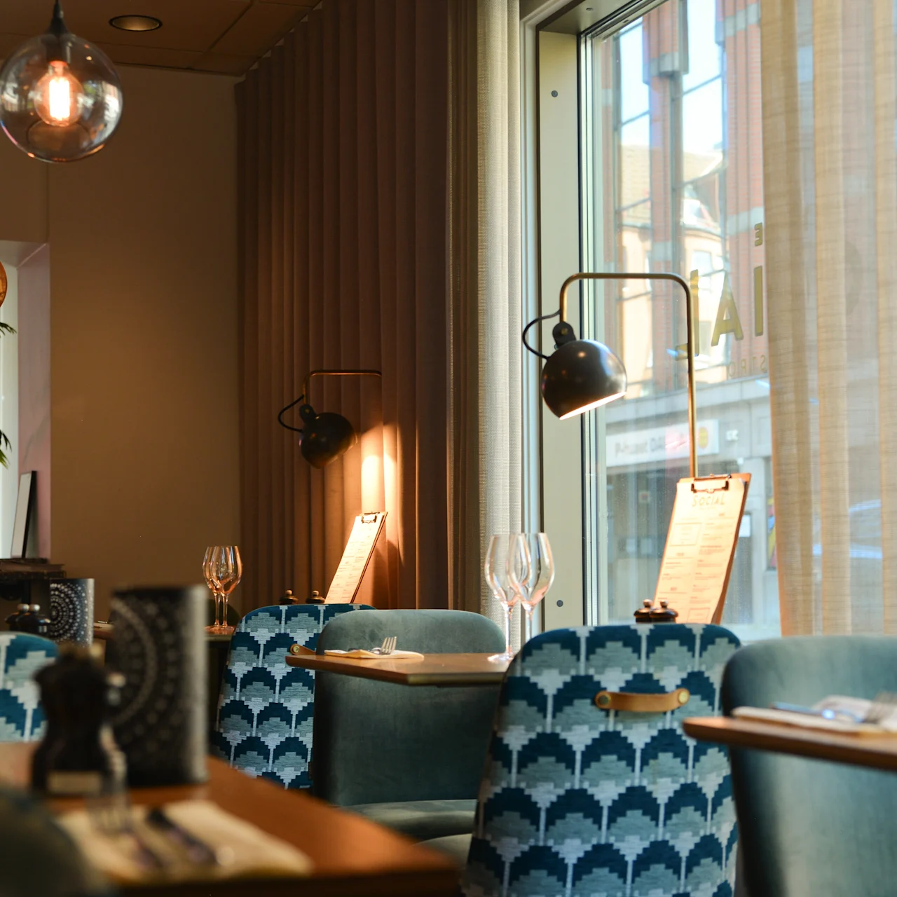 Elegant restaurant med blåmønstrede stole, oplyst af varme pendellamper, nær store vinduer. Bordene er dækket med vinglas og menuer under naturligt og omgivende lys.
