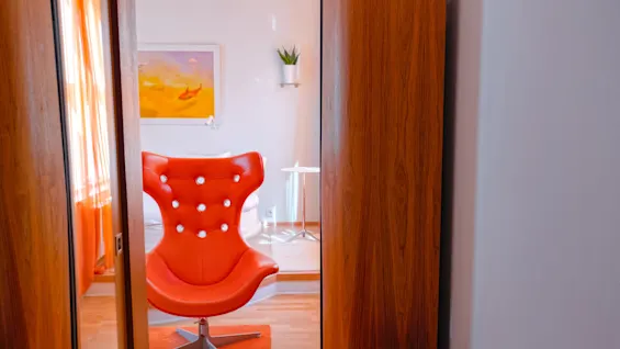 A modern, bright orange chair stands on a matching rug, framed by wooden doors, in a room with a painting, window curtains, and a potted plant on a shelf.