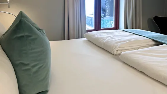 Et komfortabelt hotellrom med en seng redd opp med hvitt sengetøy og en grønn pute, ved siden av et vindu med gardiner.