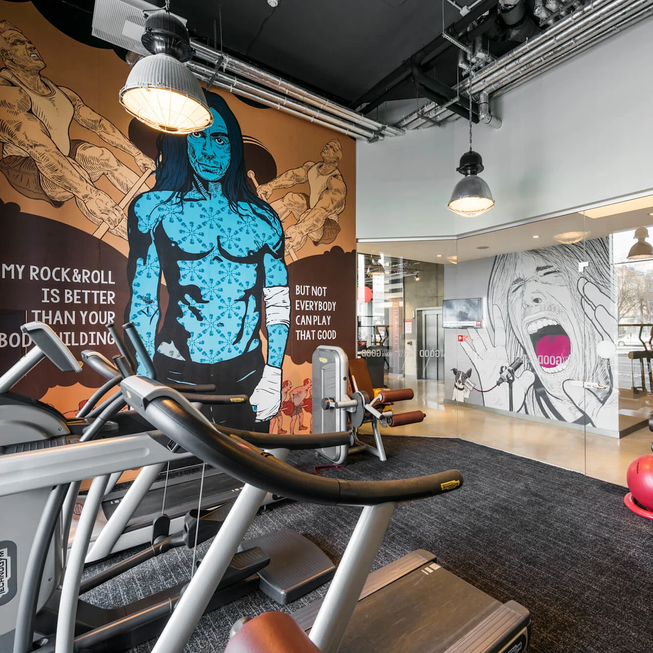 Löpband och träningsmaskiner på ett gym med målningar, inklusive en blå figur och människor som lyfter vikter. Text: "MY ROCK & ROLL IS BETTER THAN YOUR BODYBUILDING BUT NOT EVERYBODY CAN PLAY THAT GOOD."