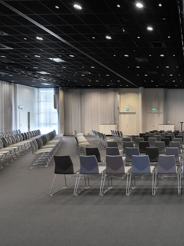 Rækker af tomme konference-stole vender mod en scene med et podium, i et stort rum med højt til loftet og grå gardiner.