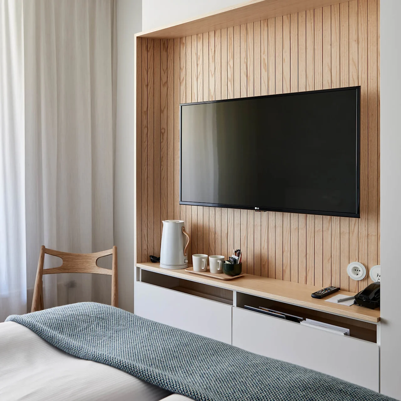 A wall-mounted TV in a bedroom above a wooden shelf holds a kettle, mugs, and remote. White curtains and a wooden chair create a minimalist, cozy atmosphere.