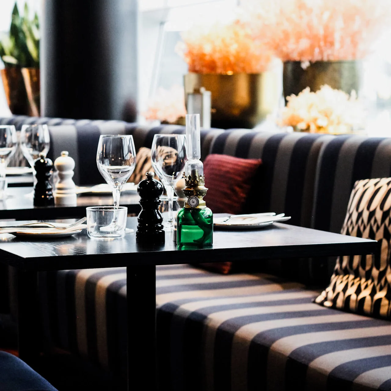 Borden är dukade med vinglas, tallrikar och bestick i en stilren och mönstrad restaurang. Soffbänkar med prydnadskuddar. Krukväxter och mjuk belysning skapar en mysig atmosfär.