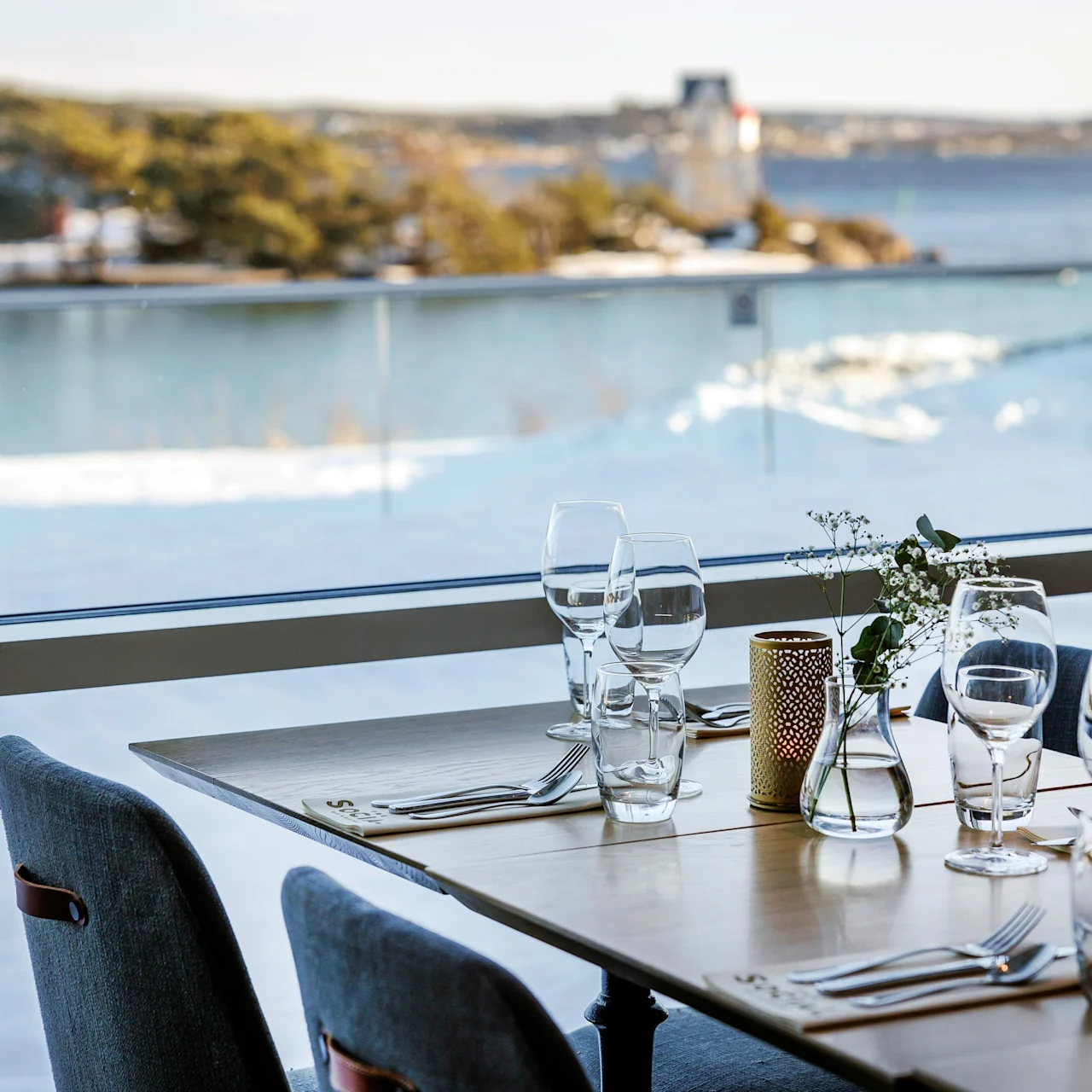 A wooden table set for dining with glassware and cutlery overlooks a snowy landscape and a body of water through a large window.