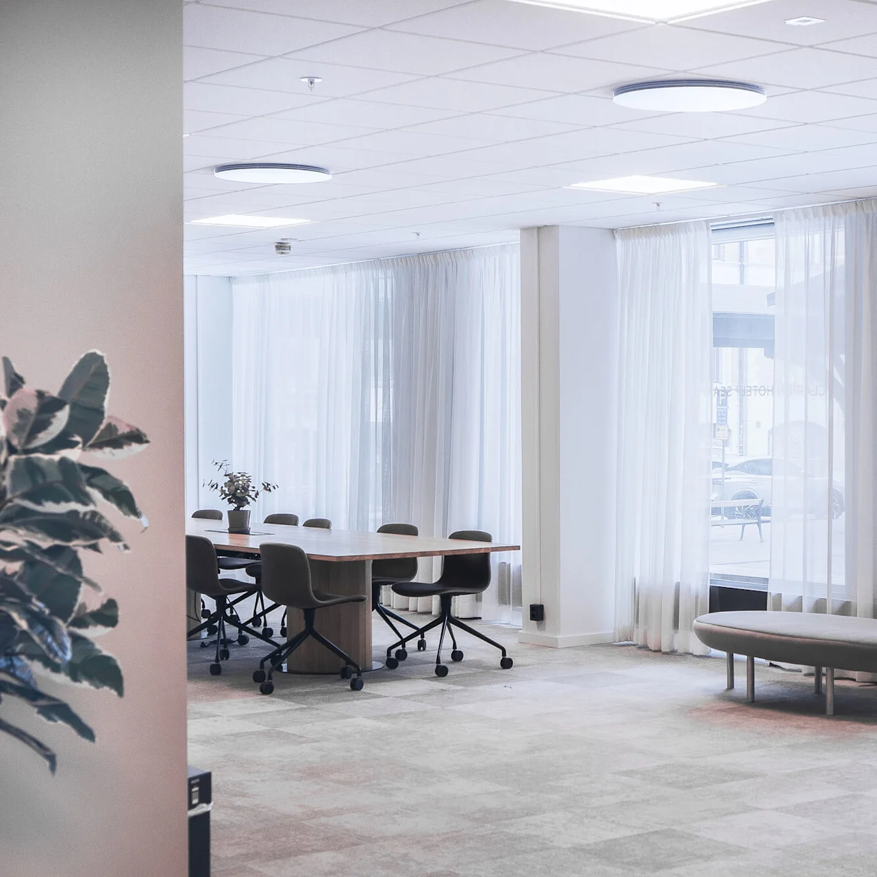 Et konferansebord er omgitt av stoler i et lyst rom med store vinduer og tynne gardiner. En potteplante står på bordet, og utenfor er biler synlige.