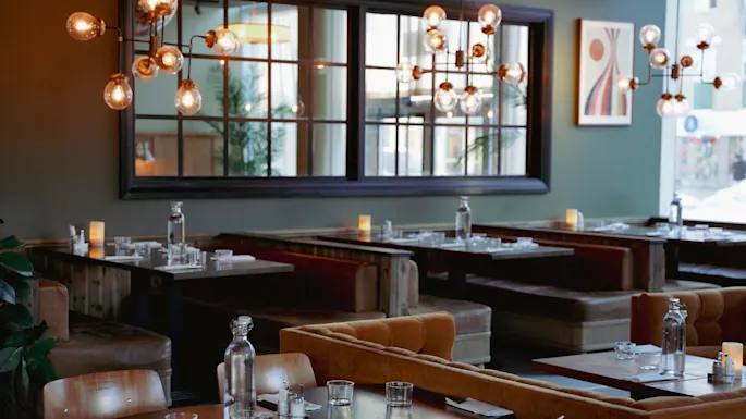 Et tomt restaurantinteriør med oppdekkede bord, hengende sfæriske lys, en grønn vegg og et vindu som viser en snødekket gate utenfor.