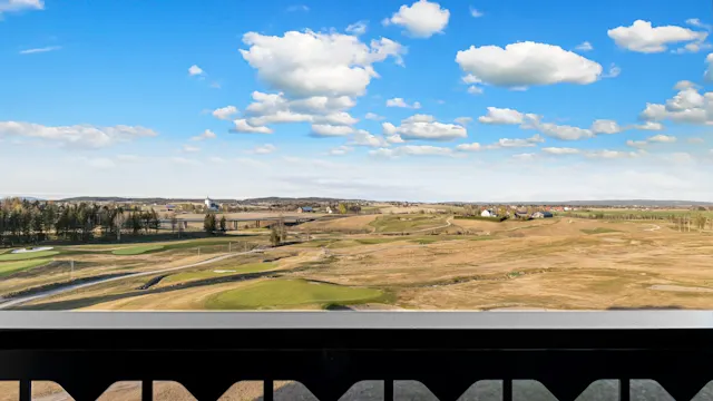 Et svart rekkverk ser ut over et vidstrakt, åpent landskap med grønne og brune jorder under en blå himmel med spredte hvite skyer. Hus er synlige i det fjerne.
