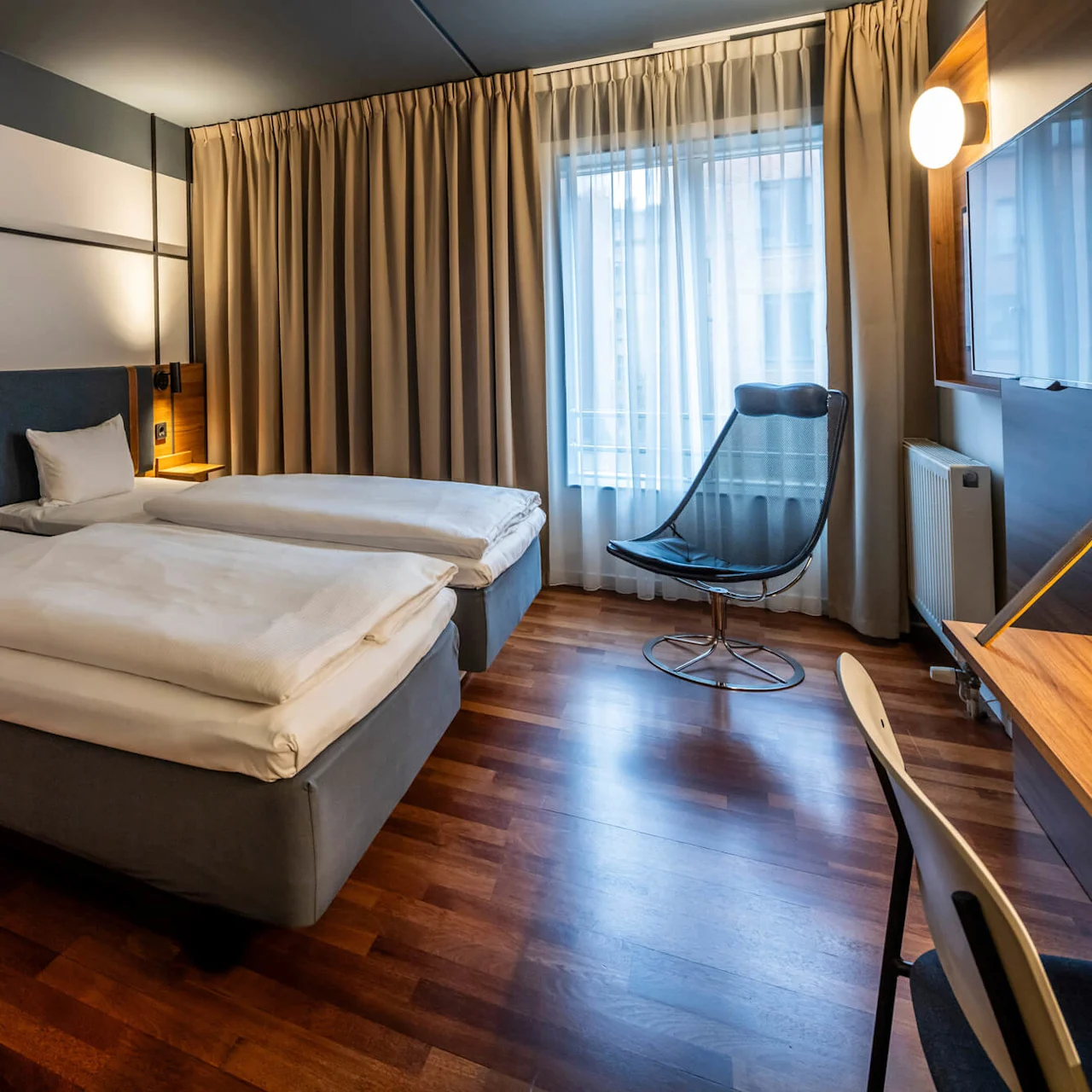 A hotel room with two single beds covered with white bedding, separated by a bedside table with a reading lamp. The room features large windows with beige curtains, a sleek chair, and a wooden desk with a minimalist chair.