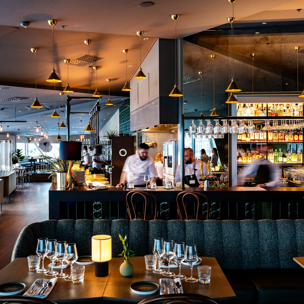 A modern restaurant features set tables with glassware and a lively bar area where staff serve drinks. The elegant lighting and stylish decor create a warm, inviting atmosphere.