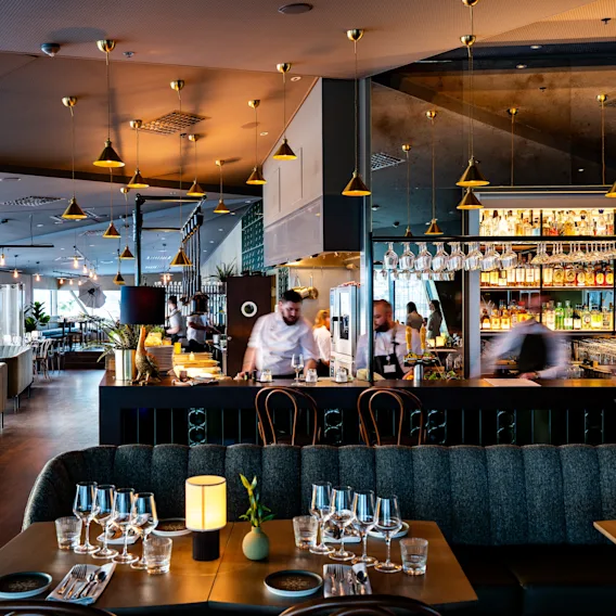 Et moderne restaurantområde med dekkede bord og glass samt en livlig bar der personalet serverer drikke. Elegant belysning og stilig innredning skaper en varm, innbydende atmosfære.