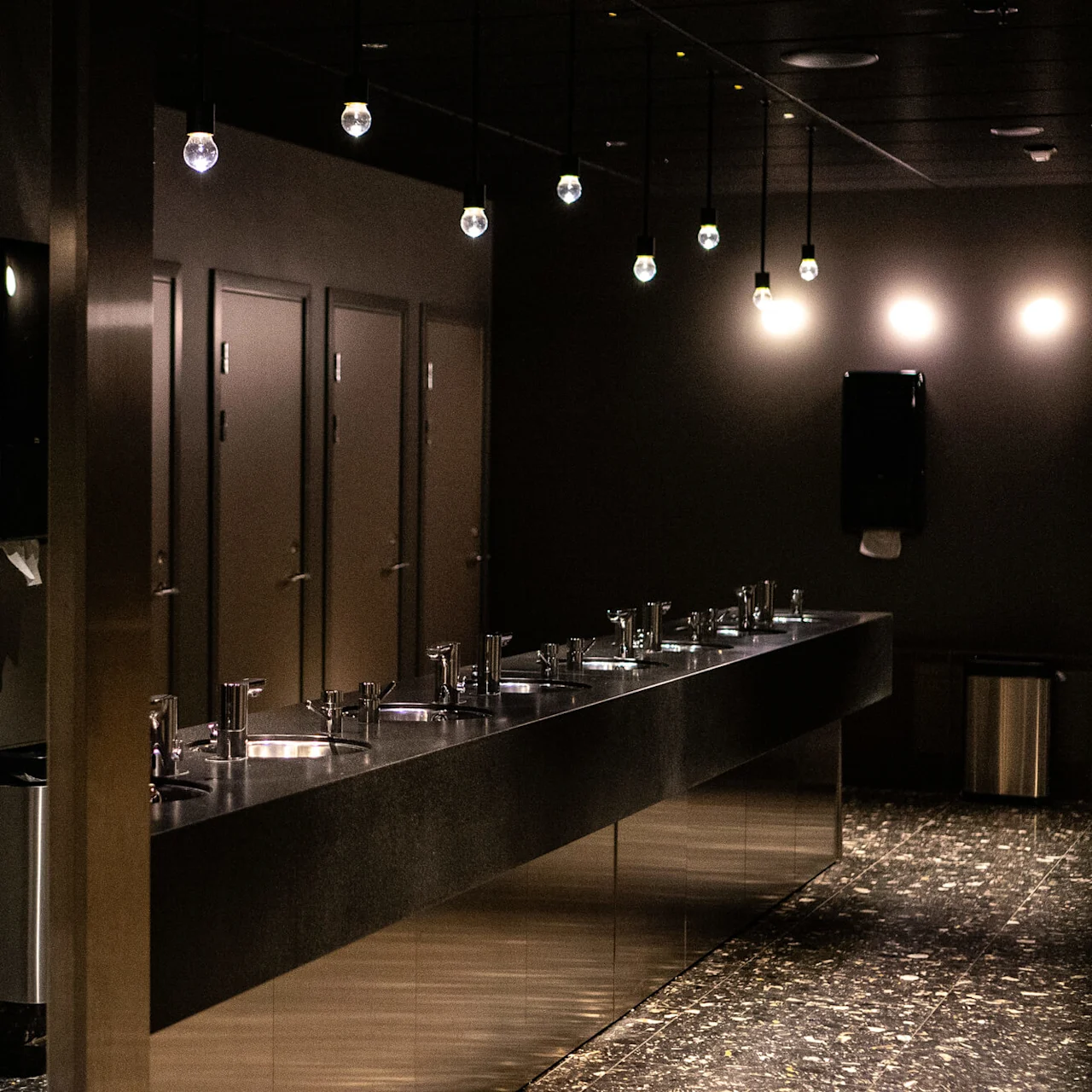 Toaletthåndvasker står på en slank benk under eksponerte lyspærer, med lukkede dører og mørke vegger som forsterker den moderne, minimalistiske atmosfæren.
