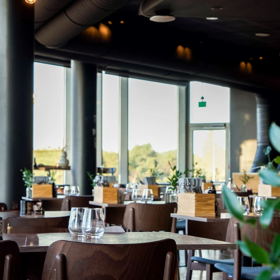 Tables with glasses are set in a modern restaurant, featuring large windows that overlook greenery outside.