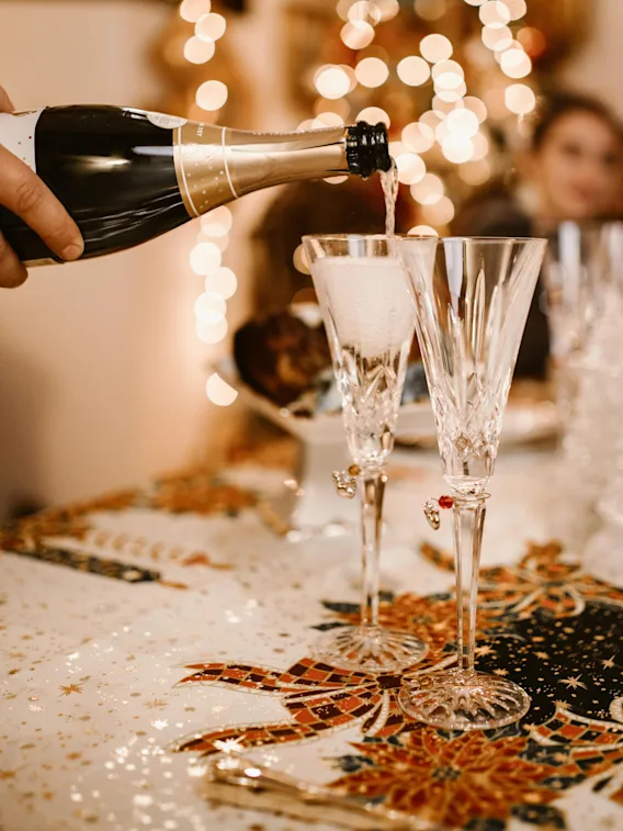 Champagne hälls i två kristallglas på ett festligt dekorerat bord med blomsterdetaljer och glittrande prydnader, i en varmt upplyst miljö med suddiga ljus i bakgrunden.