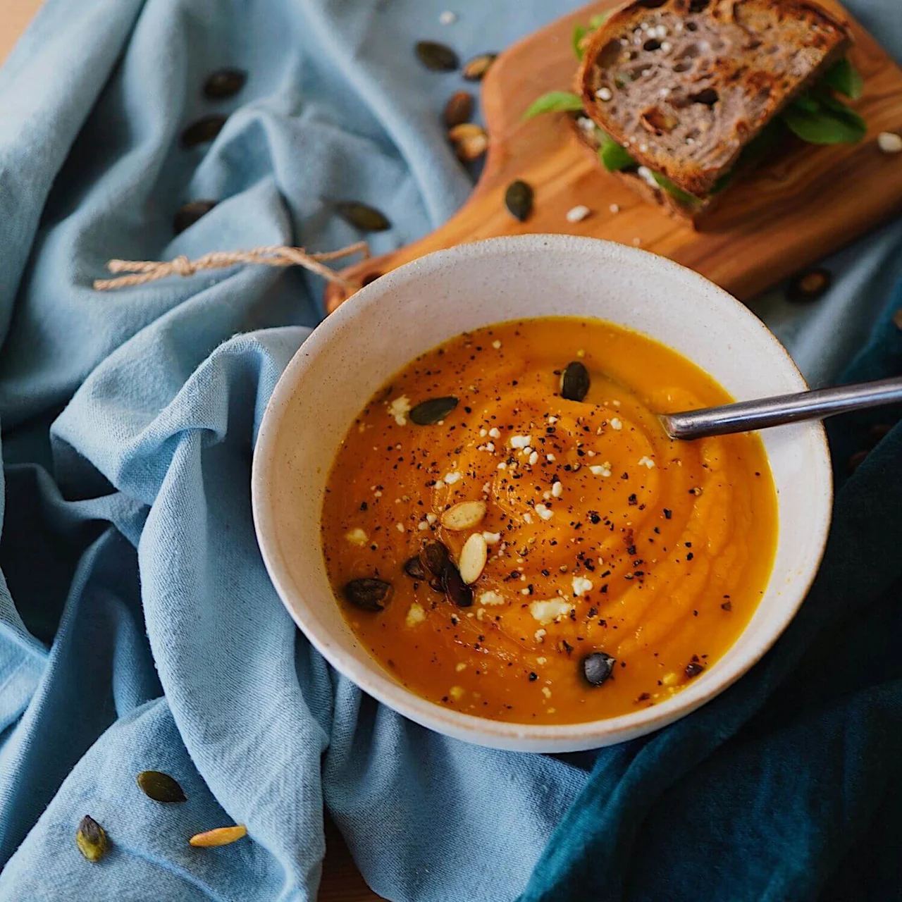 En skål med græskarsuppe pyntet med frø og krydder står på et blåt klæde med en metalske. Et træbræt har en sandwich i baggrunden.