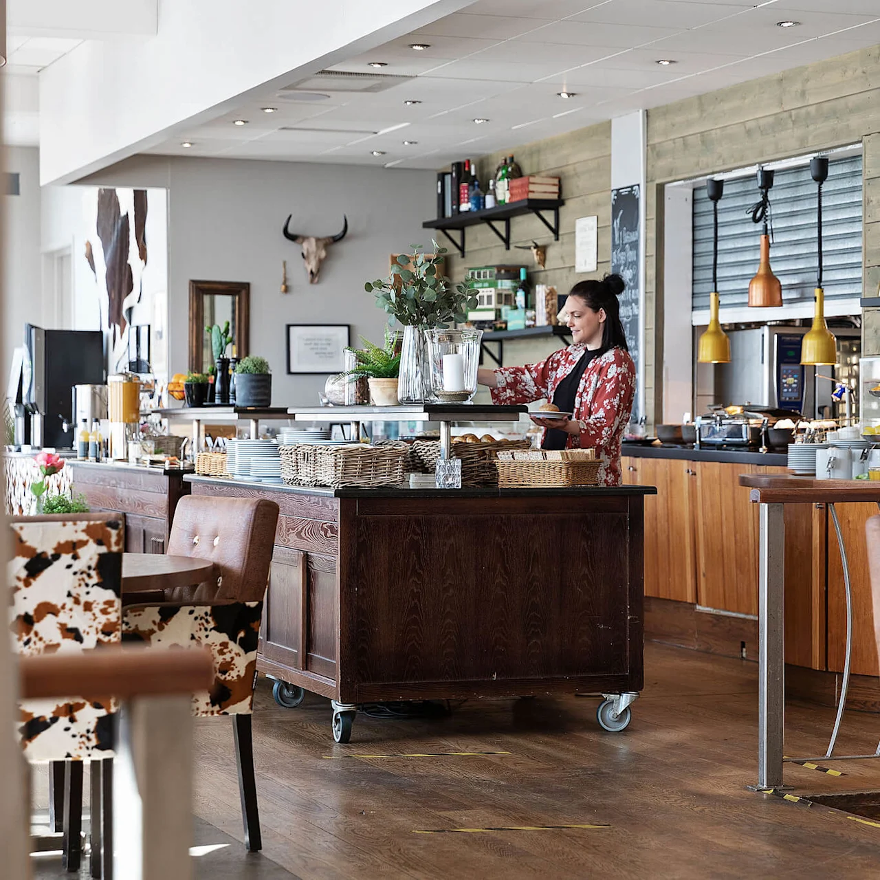 En kvinna ordnar föremål på en serveringsvagn i en restaurang omgiven av bord och en bardisk med mat och dryck.