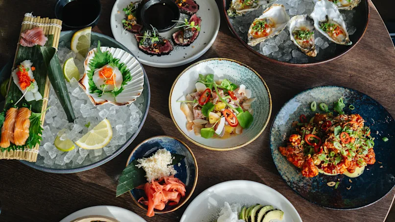 A variety of vibrant seafood dishes are artistically arranged on a wooden table. Sashimi, oysters, and ceviche are garnished with herbs, sliced chilies, and lemon wedges, surrounded by bowls and plates.