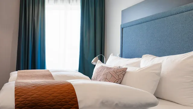 A neatly made bed with white pillows and a brown quilted runner; positioned near a large window with green curtains, creating a cozy ambiance.