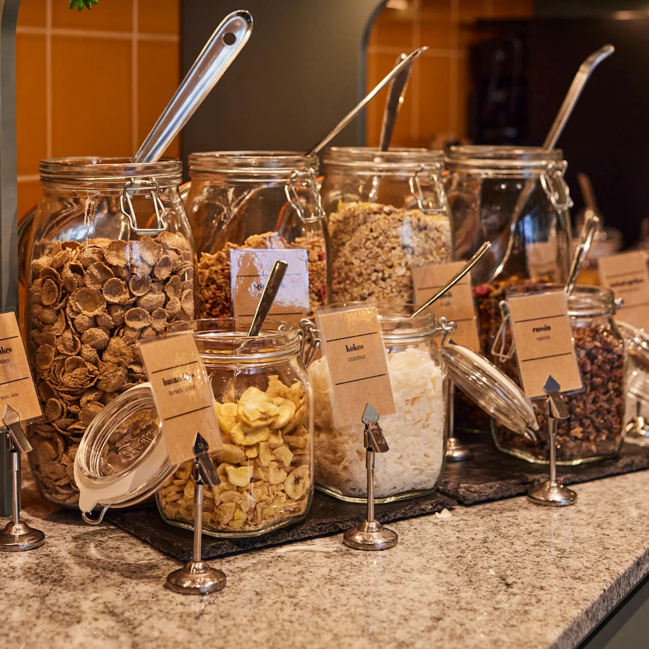 Glasskrukker fylt med ulike frokostblandinger på en kjøkkenbenk med oransje fliser og grønne planter. Etikettene lyder: "bananskivor banana chips," "kokos coconut," og "rosin raisins."