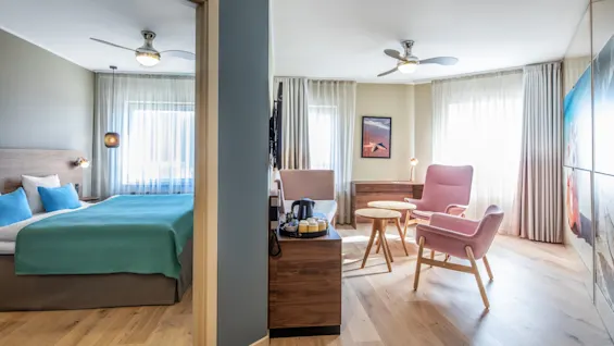 A modern hotel room features a bed with teal bedding to the left, while two pink chairs face a round table on the right, amidst bright natural light and wooden floors.