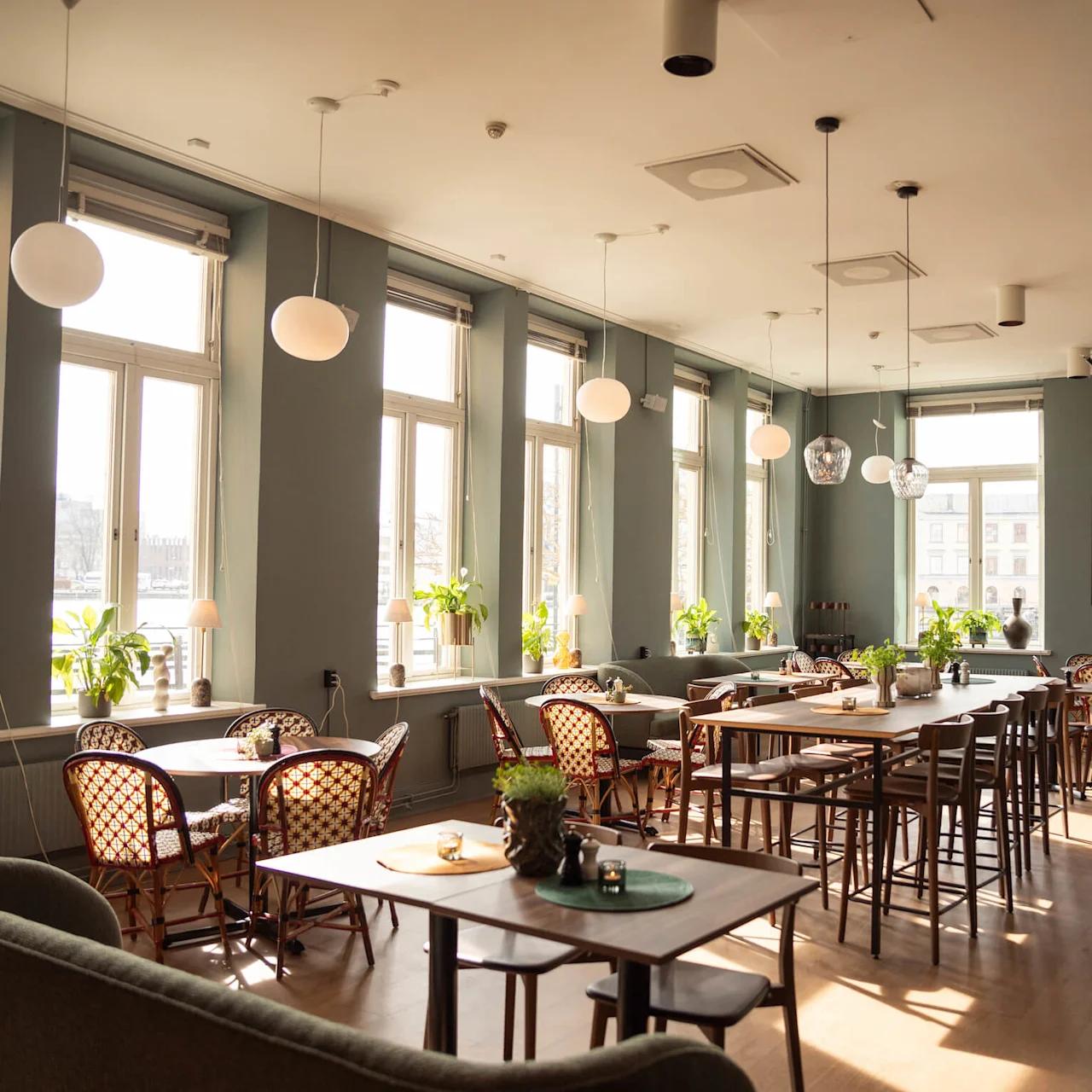Ett solbelyst café med träbord och stolar, omgiven av stora fönster och inomhusväxter, skapar en mysig och inbjudande atmosfär.