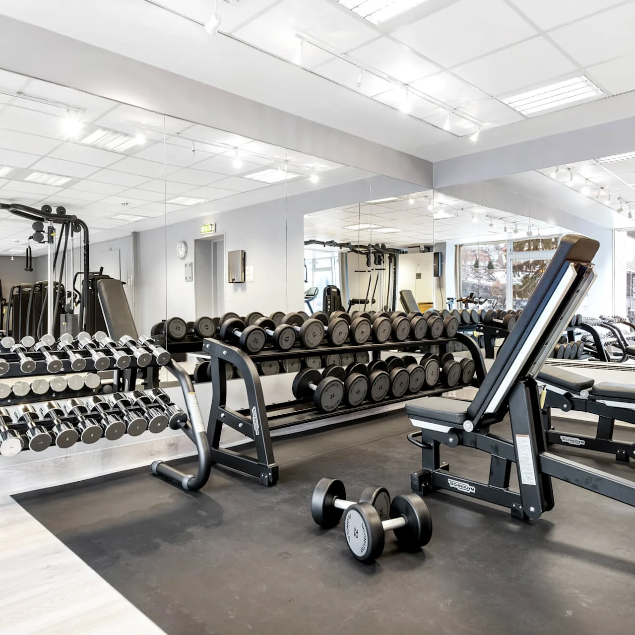 En gym har prydligt ordnade hantlar på ställ och justerbara bänkar på en svart matta, med spegelväggar som reflekterar utrustningen i en välbelyst, modern interiör.