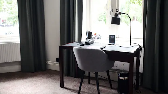 Et skrivebord med en lampe, telefon og vannkoker står ved et vindu med gardiner, noe som antyder en innstilling på et hotellrom.