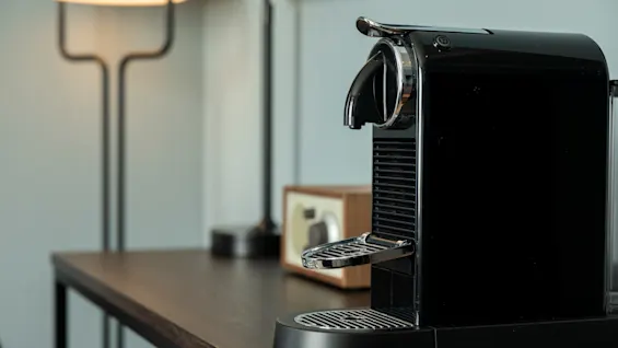 En elegant svart kaffemaskin står på et mørkt trebord, med en myk lampe og en treradio i den uskarpe bakgrunnen.