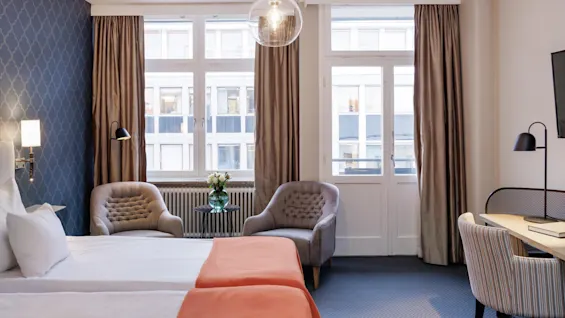 Ett välupplyst, modernt hotellrum med elegant möblering och stora fönster som erbjuder utsikt över närliggande byggnader.