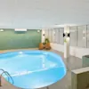 Swimmingpool – roligt vand i lyst indendørs miljø, omgivet af liggestole og potteplanter; tilstødende boblebad på flisebelagt platform.