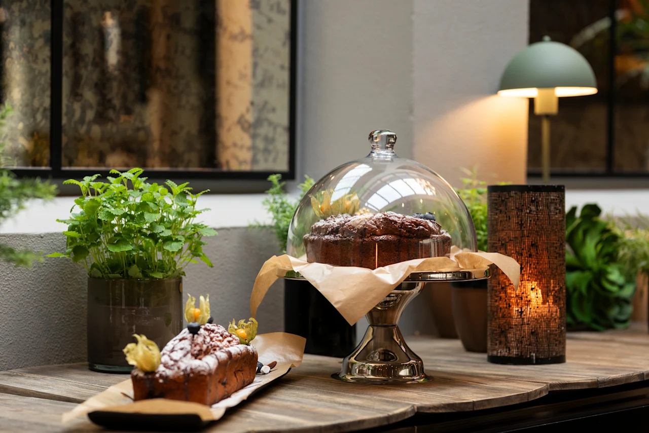Delicious cakes and pastries displayed on a wooden table with green plants and warm lighting at Home Hotel Grand Bodø.