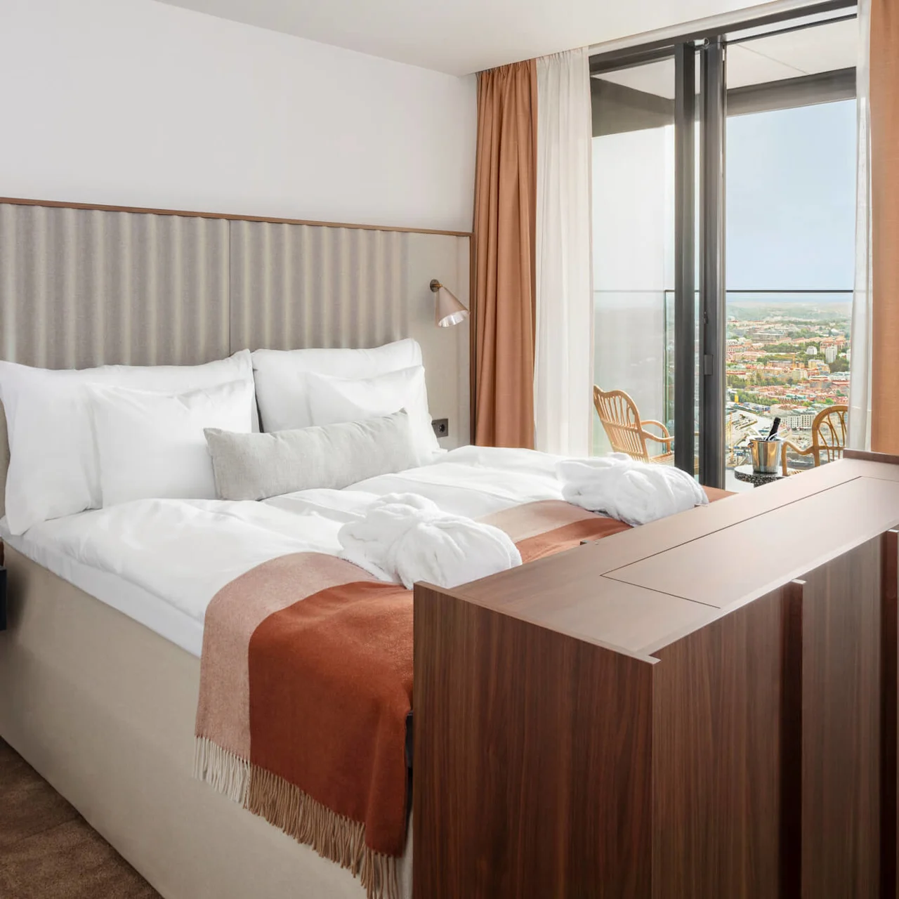 En prydligt bäddad säng med vita kuddar och en rostfärgad filt står i ett modernt hotellrum. Fönster från golv till tak avslöjar en stadsbild och två stolar utanför.